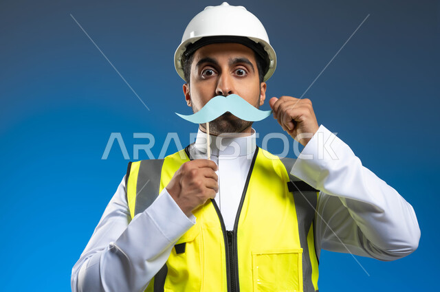 ايماءات يدين تدل على الحفاظ على القوة والصحة الجسدية، العمل في مجال الهندسة والانشاءات المعمارية، بورتريه مقرب لمهندس عربي خليجي سعودي يرتدي السترة وخوذة الحماية ويمسك عود خشبي مع قصاصة ورقية على شكل شارب، الإحتفال السنوي باليوم العالمي للرجل، خلفية زرقاء