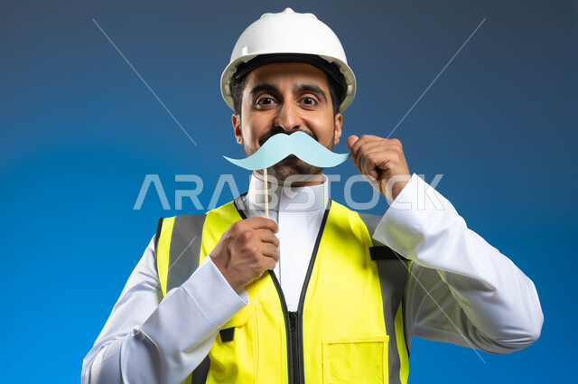 Working in the field of engineering and architectural construction, a close-up portrait of a Saudi Gulf Arab engineer wearing a jacket and a protection helmet and holding a wooden stick with a piece of paper in the shape of a mustache, hand gestures indicating the preservation of strength and physical health, the annual celebration of International Men’s Day, blue background