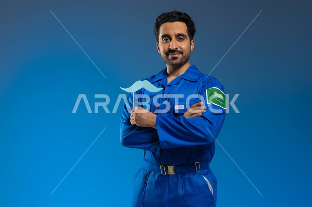 ذكرى اليوم العالمي للرجل، ايماءات النظر الى الكاميرا بأيدي متقاطعة، اتقان قيادة السفن والمركبات الفضائية، بورتريه مقرب لرائد فضاء عربي خليجي سعودي يرتدي بدلة الفضاء الزرقاء بشعار علم السعودية يحمل بيده عود خشبي مع قصاصة ورقية على شكل شارب، خلفية زرقاء