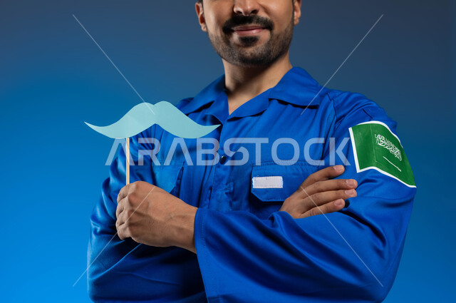 ايماءات النظر الى الكاميرا بأيدي متقاطعة، اتقان قيادة السفن والمركبات الفضائية، سنوية اليوم العالمي للرجل، بورتريه مقرب لرائد فضاء عربي خليجي سعودي يرتدي بدلة الفضاء الزرقاء بشعار علم السعودية يحمل بيده عود خشبي مع قصاصة ورقية على شكل شارب، خلفية زرقاء