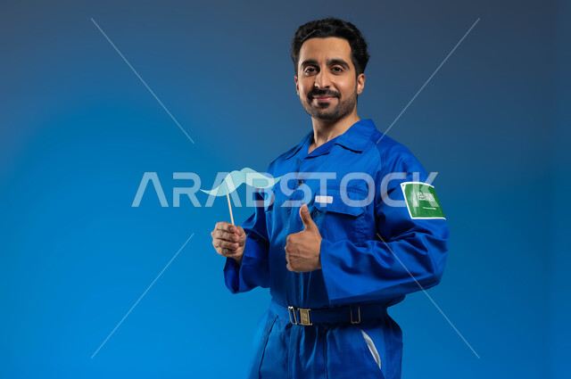 اتقان قيادة السفن والمركبات الفضائية، الاحتفال باليوم العالمي للرجل، ايماءات القبول والإعجاب بشيء ما، بورتريه مقرب لرائد فضاء عربي خليجي سعودي يرتدي بدلة الفضاء الزرقاء بشعار علم السعودية يحمل بيده عود خشبي مع قصاصة ورقية على شكل شارب، خلفية زرقاء