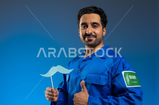 احتراف قيادة السفن والمركبات الفضائية، الاحتفال باليوم العالمي للرجل، ايماءات القبول والإعجاب بشيء ما، بورتريه مقرب لرائد فضاء عربي خليجي سعودي يرتدي بدلة الفضاء الزرقاء بشعار علم السعودية يحمل بيده عود خشبي مع قصاصة ورقية على شكل شارب، خلفية زرقاء