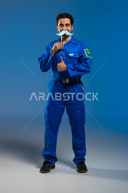 اليوم العالمي للرجل 19 نوفمبر، هندسة الفضاء والملاحة الجوية، ايماءات الإعجاب والموافقة على شيء ما، بورتريه لرائد فضاء عربي خليجي سعودي يرتدي بدلة الفضاء الزرقاء بشعار علم السعودية يحمل بيده عود خشبي مع قصاصة ورقية على شكل شارب، الطول الكامل للجسم، خلفية زرقاء