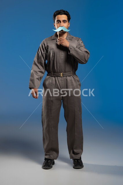 الوقوف بإستقامة والنظر الى الكاميرا، الاحتفال باليوم العالمي للرجل، مهن ووظائف وأعمال في السعودية، بورتريه لعامل صيانة عربي خليجي سعودي يرتدي الزي الخاص بالعمل يحمل بيده عود خشبي مع قصاصة ورقية على شكل شارب يغطي فمه بها، الطول الكامل للجسم، خلفية زرقاء