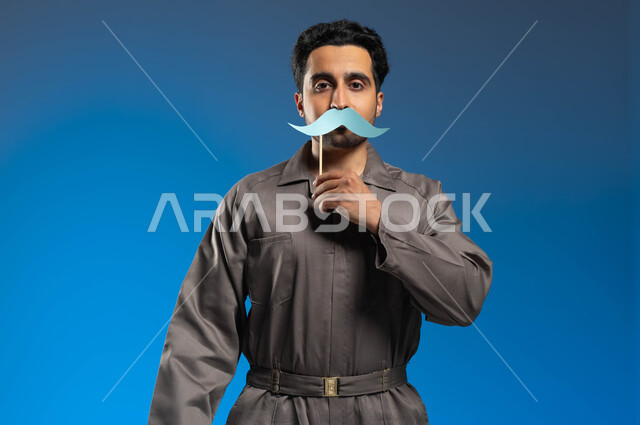 الاحتفال باليوم العالمي للرجل، الوقوف بإستقامة والنظر الى الكاميرا، مهن ووظائف وأعمال في السعودية، بورتريه مقرب لعامل صيانة عربي خليجي سعودي يرتدي الزي الخاص بالعمل يحمل بيده عود خشبي مع قصاصة ورقية على شكل شارب يغطي فمه بها، خلفية زرقاء