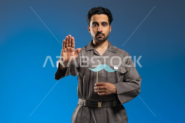 ايماءات الإنزعاج ورفع اليد للحذر من الاقتراب، مفهوم الأعمال والصيانات في السعودية، إحياء ذكرى اليوم العالمي للرجل 19 نوفمبر، بورتريه مقرب لعامل صيانة عربي خليجي سعودي يرتدي الزي الخاص بالعمل ويحمل بيده عود خشبي مع قصاصة ورقية على شكل شارب، خلفية زرقاء