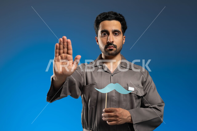 إحياء ذكرى اليوم العالمي للرجل 19 نوفمبر، ايماءات الإنزعاج ورفع اليد للحذر من الاقتراب، مفهوم الأعمال والصيانات في السعودية، بورتريه مقرب لعامل صيانة عربي خليجي سعودي يرتدي الزي الخاص بالعمل ويحمل بيده عود خشبي مع قصاصة ورقية على شكل شارب، خلفية زرقاء