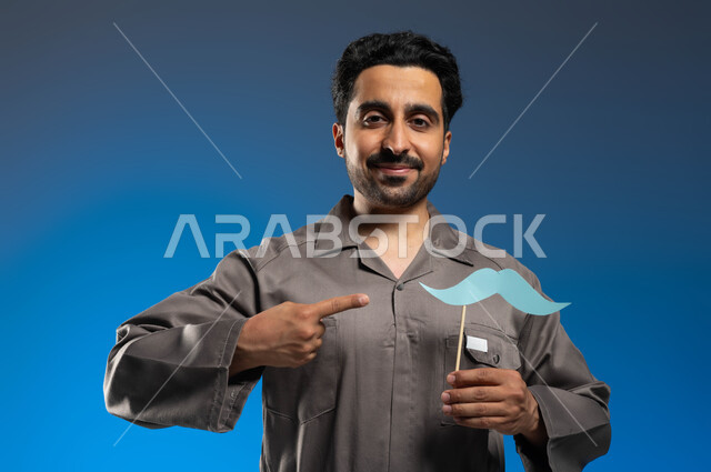 النظر الى الكاميرا بايماءات السرور والثقة بالنفس، إتقان مهنة فني الصيانة بإحتراف، ذكرى اليوم العالمي للرجل 19 نوفمبر، بورتريه مقرب لعامل صيانة عربي خليجي سعودي يرتدي الزي الخاص بالعمل يشير بإصبعه الى عود خشبي مع قصاصة ورقية على شكل شارب، خلفية زرقاء