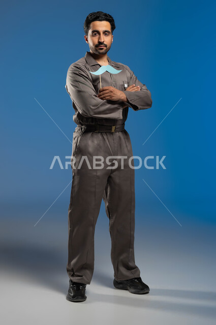 النظر الى الكاميرا بإبتسامة وايدي متقاطعة، مزاولة مهنة فني الصيانة بإحتراف، ذكرى اليوم العالمي للرجل 19 نوفمبر، بورتريه لعامل صيانة عربي خليجي سعودي يرتدي الزي الخاص بالعمل ويحمل بيده عود خشبي مع قصاصة ورقية على شكل شارب، الطول الكامل للجسم، خلفية زرقاء