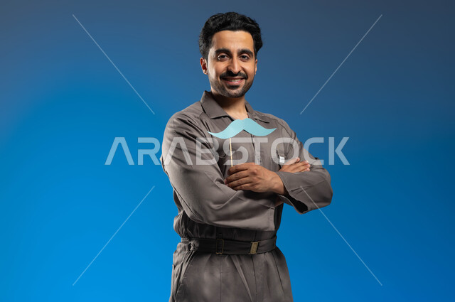 مزاولة مهنة فني الصيانة بإحتراف، ذكرى اليوم العالمي للرجل 19 نوفمبر، بورتريه مقرب لعامل صيانة عربي خليجي سعودي يرتدي الزي الخاص بالعمل ويحمل بيده عود خشبي مع قصاصة ورقية على شكل شارب، النظر الى الكاميرا بإبتسامة وايدي متقاطعة، خلفية زرقاء