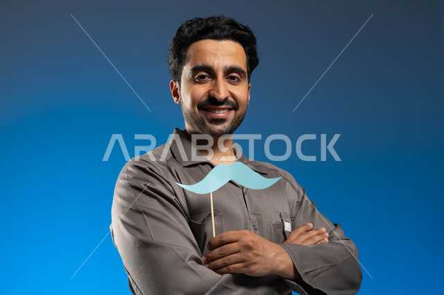 ذكرى اليوم العالمي للرجل 19 نوفمبر، بورتريه مقرب لعامل صيانة عربي خليجي سعودي يرتدي الزي الخاص بالعمل ويحمل بيده عود خشبي مع قصاصة ورقية على شكل شارب، مزاولة مهنة فني الصيانة بإحتراف، النظر الى الكاميرا بإبتسامة وايدي متقاطعة، خلفية زرقاء