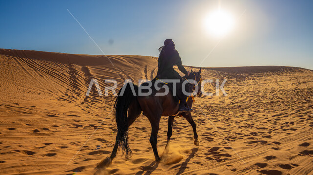 Enjoying practicing traditional hobbies, strolling on the soft golden sand in one of the Kingdom’s deserts, a picture from behind of a Saudi Arabian Gulf horsewoman wearing a black abaya riding a purebred brown mare, Our Day 1727 AD, the anniversary of the founding of the first Saudi state on February 22
