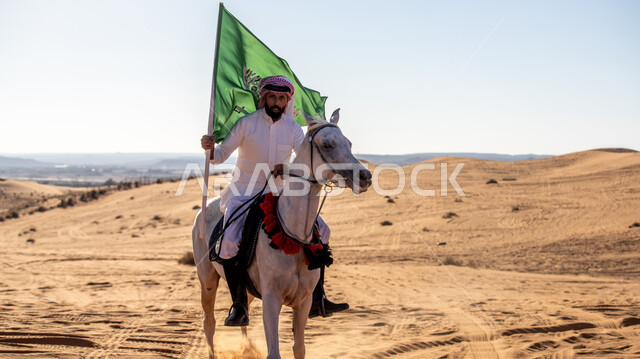Practicing the hobby of horse riding in the desert nature, a Saudi Gulf Arab knight wearing the traditional dress and shemagh, riding a purebred horse and holding the Kingdom’s flag proudly in his hand, Day of Our Day 1727 AD, the founding of the first Saudi state on February 22, commemorating the National Day on September 23, Flag Day on March 11