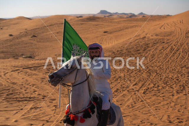 Practicing the hobby of horse riding in the desert nature, a Saudi Gulf Arab knight wearing the traditional dress and shemagh, riding a purebred horse and holding the Kingdom’s flag proudly in his hand, Day of Our Day 1727 AD, the founding of the first Saudi state on February 22, commemorating the National Day on September 23, Flag Day on March 11