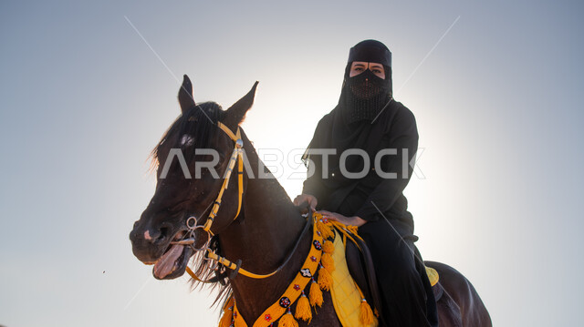 Enjoying practicing traditional hobbies, riding a horse in one of the Kingdom’s deserts, a Saudi Arabian Gulf horsewoman wearing a burqa and black abaya riding a purebred brown horse, Day of Our Day 1727 AD, the anniversary of the founding of the first Saudi state on February 22, inheriting the love of horses from the ancestors