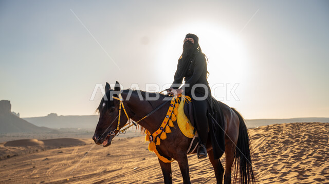 Enjoying practicing traditional hobbies, riding a horse in the Arabian desert during the day, a Saudi Arabian Gulf horsewoman wearing a burqa and black abaya riding a purebred brown horse, Day of Fatty 1727 AD, the anniversary of the founding of the first Saudi state on February 22, following the traditions of our ancestors in loving and raising horses