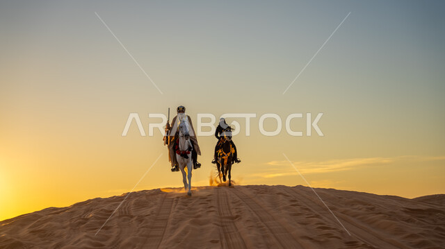 The strength and authenticity of the Arabian horse, events and activities riding Arabian horses in the desert on the occasion of the anniversary of the founding of the first Saudi state, February 22, two Saudi Arabian Gulf horsemen wearing popular traditional costumes riding horses racing over the golden sands during the day, Day of Dina 1727 AD