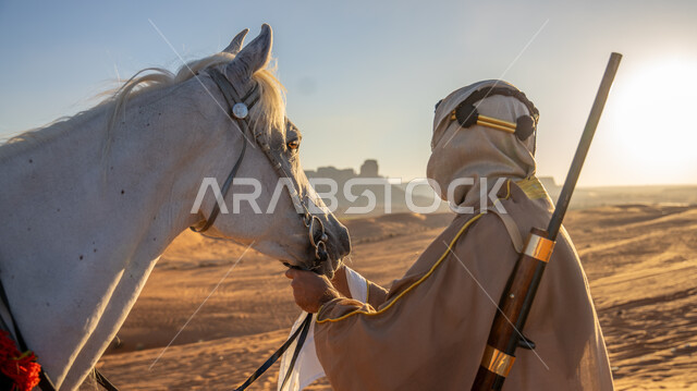 رعاية الخيل والاعتناء به في الصحراء وضح النهار، ذكرى تأسيس الدولة السعودية الأولى 22 فبراير، يوم بدينا 1727 م، اتباع تقاليد الأجداد في حب وتربية الجياد العربية، صورة من الخلف لرجل عربي خليجي سعودي يرتدي البشت والغترة مع العقال يقوم بملاطفة ومداعبة فرسه الأبيض