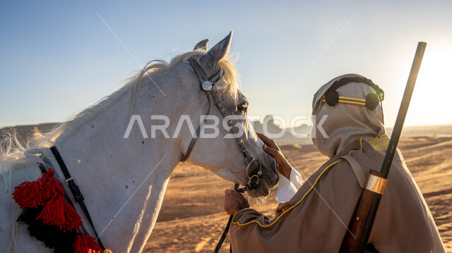  الاعتناء بالخيل ورعايته في الصحراء وضح النهار، ذكرى تأسيس الدولة السعودية الأولى 22 فبراير، يوم بدينا 1727 م، اتباع تقاليد الأجداد في حب وتربية الجياد العربية، صورة من الخلف لرجل عربي خليجي سعودي يرتدي البشت والغترة مع العقال يقوم بملاطفة ومداعبة فرسه الأبيض