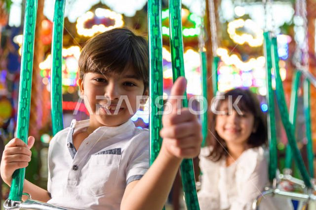 Two Saudi children in the amusement park, playing the fun game of swings, spending great times in the amusement park, electronic games, entertainment and entertainment, during the summer vacation