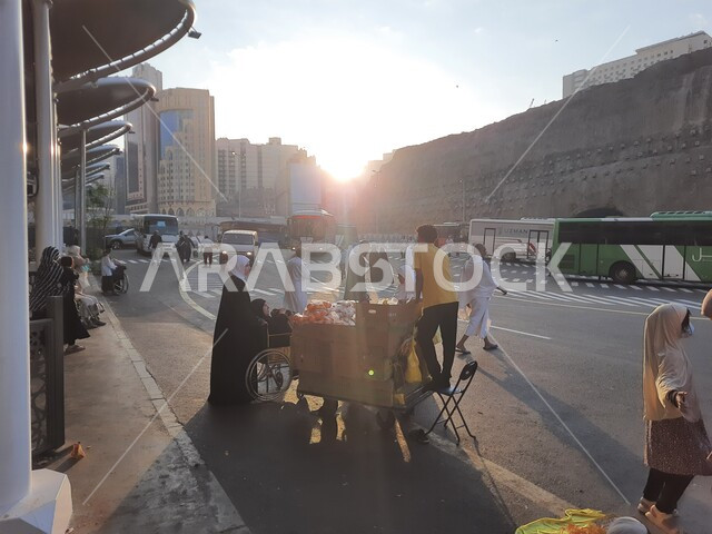مجموعة من حجاج بيت الله الحرام في موقف للحافلات بجوار الحرم المكي بمكة المكرمة وقت غروب الشمس، معالم واماكن اسلامية دينية مقدسة، العبادة والتقرب إلى الله، التجمع في ساحة للمواصلات ووسائل النقل استعدادا للتنقل
