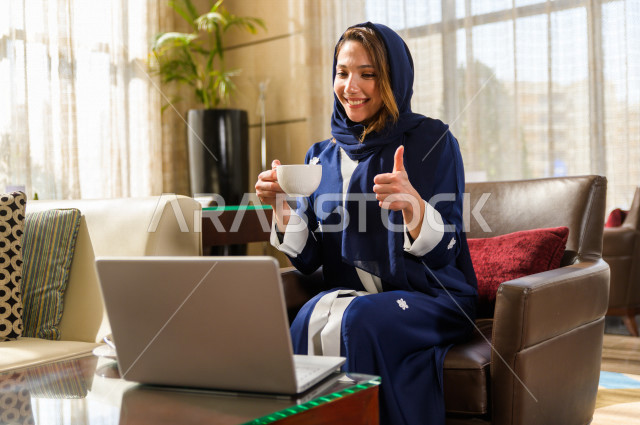 مرأه عربية خليجية سعيدة في احدى المتاجر ،تعمل على الكمبيوتر المحمول ، بجانبها  فنجان القهوة ،  متجر القهوه ، بيئة العمل