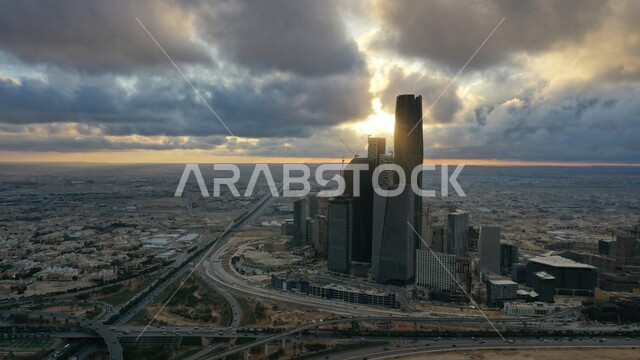 منظر الشمس وقت الغروب, السماء المليئة بالغيوم, الاشجار والنباتات الخضراء, النمو والتطور الهندسي المعماري للأبراج وناطحات السحاب بالسعودية، حركة سير السيارات على الطريق العام، مركز الملك عبدالله المالي (كافد) في العاصمة، تحقيق النهضة الاقتصادية والتجارية