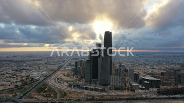 View of the sun at sunset, cloudy sky, trees and green plants, growth and architectural development of towers and skyscrapers in Saudi Arabia, car traffic on the public road, King Abdullah Financial Center (CAFD) in the capital, achieving economic and commercial renaissance