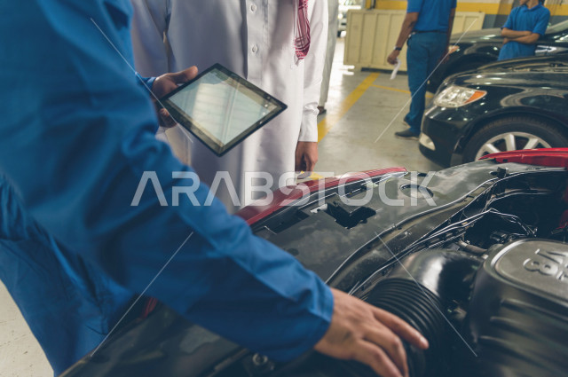 رجل عربي سعودي  داخل ورشة السيارات ، يستخدم جهاز التابلت لاختيار خدمة التصليح ، معه مهندس ميكانيكي يقوم باصلاح سيارته ، استخدام ادوات للصيانة ، اصلاح يدوي ، خدمات اصلاح و صيانة السيارات ، مهندس ميكانيكي ، ورشة صيانة 