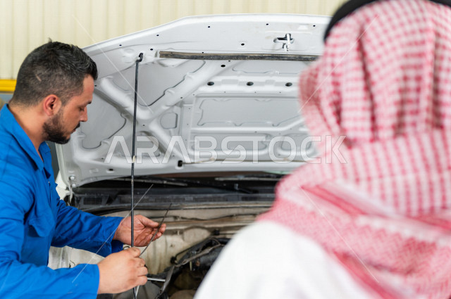 رجل عربي سعودي  داخل ورشة السيارات  ، بصحبة مهندس ميكانيكي يقوم باصلاح سيارته ، استخدام ادوات للصيانة ، اصلاح يدوي ، خدمات اصلاح و صيانة السيارات ، مهندس ميكانيكي ، ورشة صيانة 