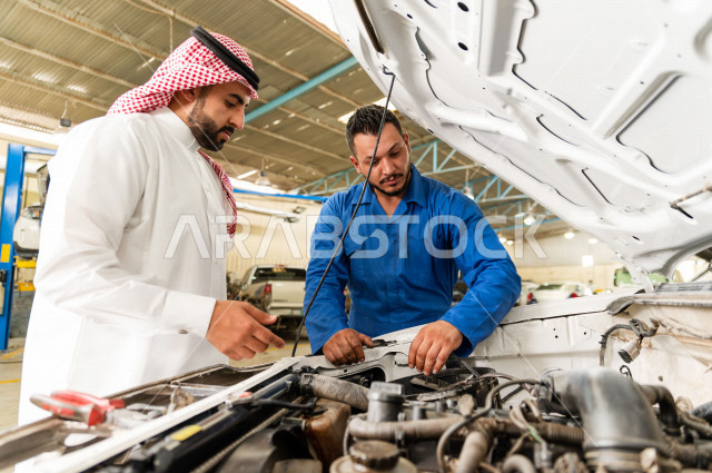 رجل عربي سعودي  داخل ورشة السيارات  ، بصحبة مهندس ميكانيكي يقوم باصلاح سيارته ، استخدام ادوات للصيانة ، اصلاح يدوي ، خدمات اصلاح و صيانة السيارات ، مهندس ميكانيكي ، ورشة صيانة 