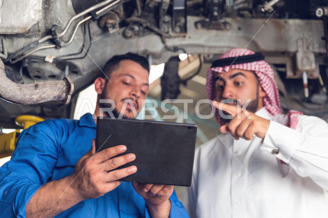 رجل عربي سعودي  داخل ورشة السيارات ، يستخدم جهاز التابلت لاختيار خدمة التصليح ، معه مهندس ميكانيكي يقوم باصلاح سيارته ، استخدام ادوات للصيانة ، اصلاح يدوي ، خدمات اصلاح و صيانة السيارات ، مهندس ميكانيكي ، ورشة صيانة 