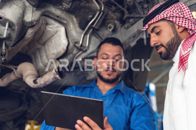 رجل عربي سعودي  داخل ورشة السيارات ، يستخدم جهاز التابلت لاختيار خدمة التصليح ، معه مهندس ميكانيكي يقوم باصلاح سيارته ، استخدام ادوات للصيانة ، اصلاح يدوي ، خدمات اصلاح و صيانة السيارات ، مهندس ميكانيكي ، ورشة صيانة 
