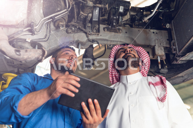 رجل عربي سعودي  داخل ورشة السيارات ، يستخدم جهاز التابلت لاختيار خدمة التصليح ، معه مهندس ميكانيكي يقوم باصلاح سيارته ، استخدام ادوات للصيانة ، اصلاح يدوي ، خدمات اصلاح و صيانة السيارات ، مهندس ميكانيكي ، ورشة صيانة 