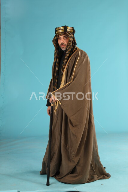 ذكرى تأسيس الدولة السعودية الأولى في 22 فبراير، يوم بدينا 1727م، بورتريه رجل عربي خليجي سعودي يرتدي البشت والعقال الدهبي ينظر إلى الكاميرا، الفخر والتباهي بانجازات البلاد، صورة الجسم بالطول الكامل بخلفية زرقاء