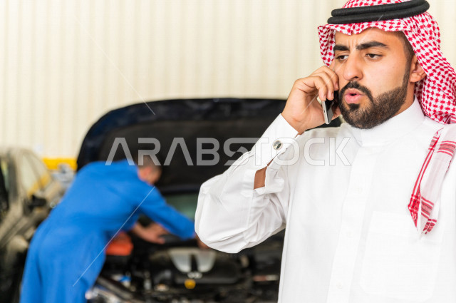 A Saudi Arabian man inside the car workshop, uses his mobile phone, online services, a mechanical engineer who repairs his car, uses tools for maintenance, manual repair, car repair and maintenance services, a mechanical engineer, a maintenance workshop