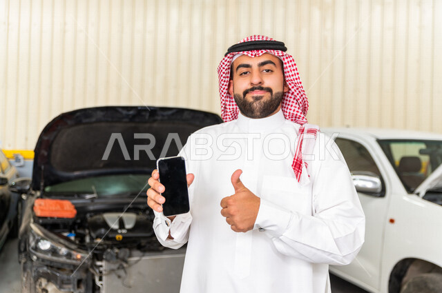 رجل عربي سعودي  داخل ورشة السيارات ، يستخدم هاتفه المحمول ، خدمات اونلاين ، مهندس ميكانيكي يقوم باصلاح سيارته ، استخدام ادوات للصيانة ، اصلاح يدوي ، خدمات اصلاح و صيانة السيارات ، مهندس ميكانيكي ، ورشة صيانة 
