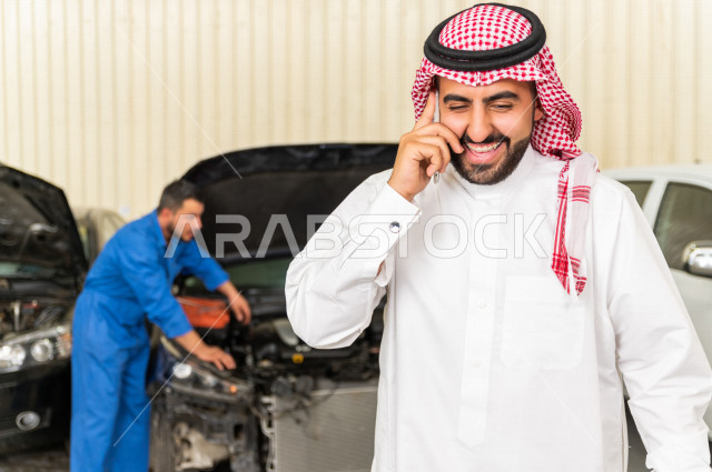 رجل عربي سعودي  داخل ورشة السيارات ، يستخدم هاتفه المحمول ، خدمات اونلاين ، مهندس ميكانيكي يقوم باصلاح سيارته ، استخدام ادوات للصيانة ، اصلاح يدوي ، خدمات اصلاح و صيانة السيارات ، مهندس ميكانيكي ، ورشة صيانة 