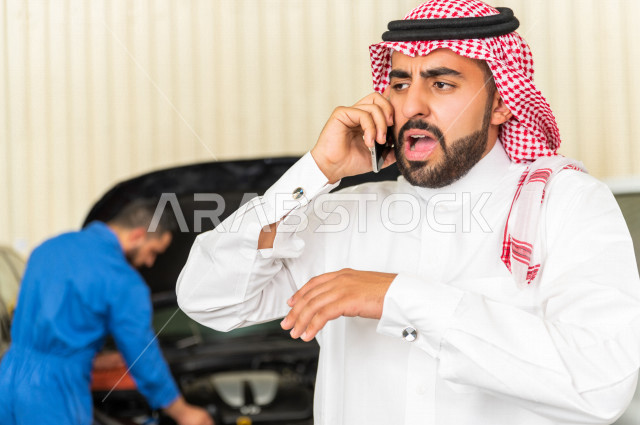 رجل عربي سعودي  داخل ورشة السيارات ، يستخدم هاتفه المحمول ، خدمات اونلاين ، مهندس ميكانيكي يقوم باصلاح سيارته ، استخدام ادوات للصيانة ، اصلاح يدوي ، خدمات اصلاح و صيانة السيارات ، مهندس ميكانيكي ، ورشة صيانة 