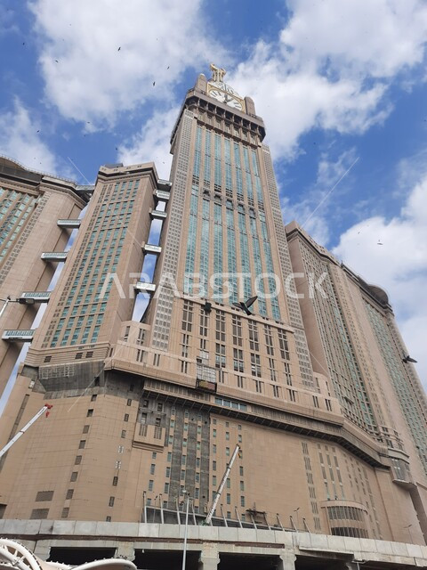 السماء المليئة بالغيوم فوق الأبراج المطلة على الحرم المكي الشريف في المملكة العربية السعودية، مبنى برج الساعة الملكي في مكة المكرمة في النهار، الفن الهندسي العمراني على الطراز الإسلامي، معالم دينية اسلامية مشهورة، بيت الله الحرام