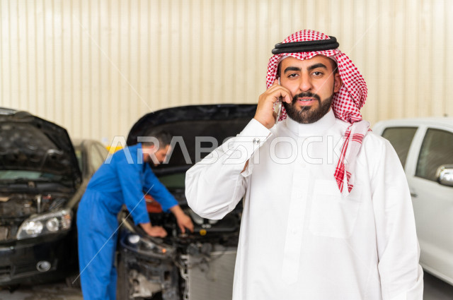 رجل عربي سعودي  داخل ورشة السيارات ، يستخدم هاتفه المحمول ، خدمات اونلاين ، مهندس ميكانيكي يقوم باصلاح سيارته ، استخدام ادوات للصيانة ، اصلاح يدوي ، خدمات اصلاح و صيانة السيارات ، مهندس ميكانيكي ، ورشة صيانة 