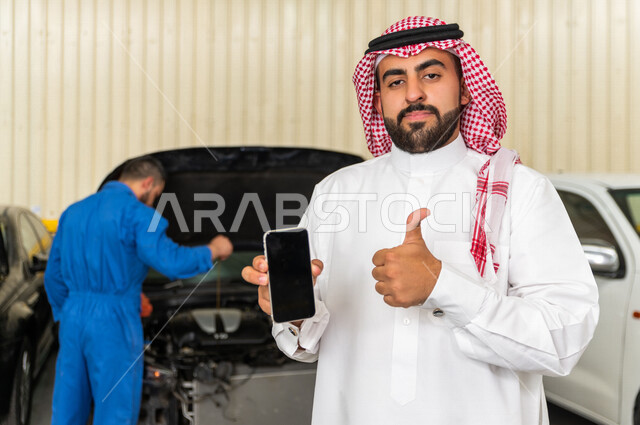 رجل عربي سعودي  داخل ورشة السيارات ، يستخدم هاتفه المحمول ، خدمات اونلاين ، مهندس ميكانيكي يقوم باصلاح سيارته ، استخدام ادوات للصيانة ، اصلاح يدوي ، خدمات اصلاح و صيانة السيارات ، مهندس ميكانيكي ، ورشة صيانة 