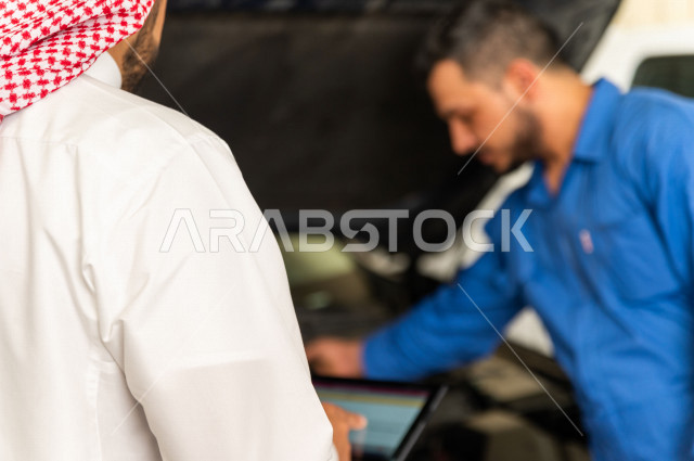 رجل عربي سعودي  داخل ورشة السيارات ، يستخدم جهاز التابلت لاختيار خدمة التصليح ، معه مهندس ميكانيكي يقوم باصلاح سيارته ، استخدام ادوات للصيانة ، اصلاح يدوي ، خدمات اصلاح و صيانة السيارات ، مهندس ميكانيكي ، ورشة صيانة 