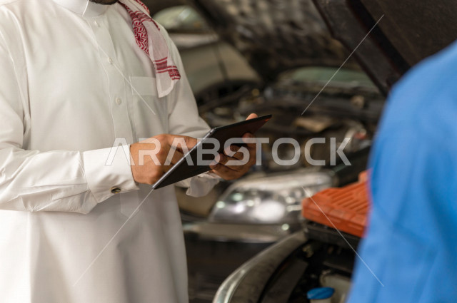 رجل عربي سعودي  داخل ورشة السيارات ، يستخدم جهاز التابلت لاختيار خدمة التصليح ، معه مهندس ميكانيكي يقوم باصلاح سيارته ، استخدام ادوات للصيانة ، اصلاح يدوي ، خدمات اصلاح و صيانة السيارات ، مهندس ميكانيكي ، ورشة صيانة 