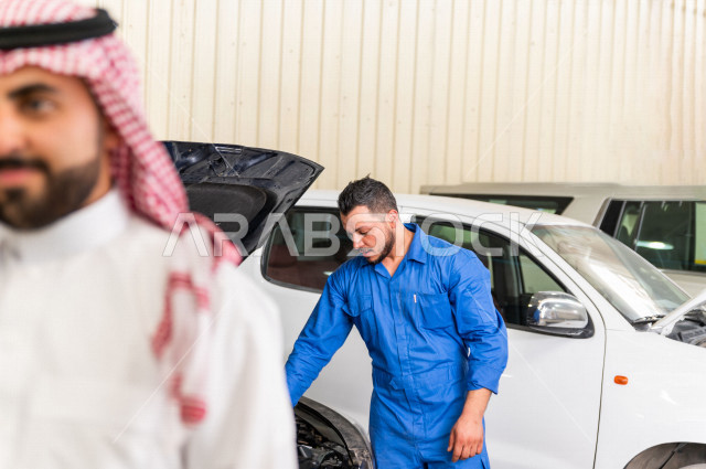 رجل  عربي سعودي يقف مع المهندس الميكانيكي داخل ورشة السيارات  ، بايماءات وجه و يدين مختلفة  ، خدمات اصلاح و صيانة السيارات ، مهندس ميكانيكي ، ورشة صيانة 