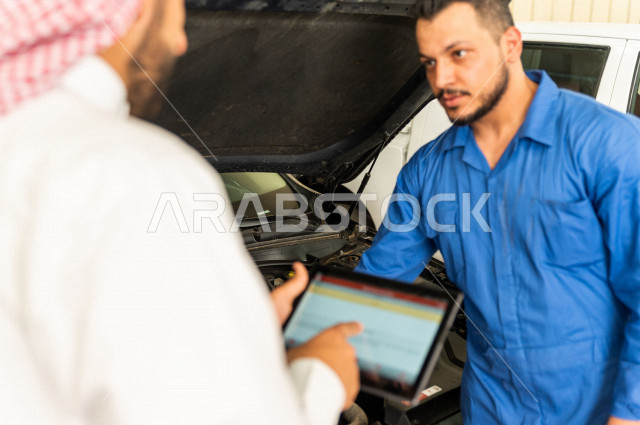 رجل عربي سعودي  داخل ورشة السيارات ، يستخدم جهاز التابلت لاختيار خدمة التصليح ، معه مهندس ميكانيكي يقوم باصلاح سيارته ، استخدام ادوات للصيانة ، اصلاح يدوي ، خدمات اصلاح و صيانة السيارات ، مهندس ميكانيكي ، ورشة صيانة 