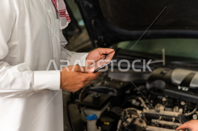 رجل عربي سعودي  داخل ورشة السيارات ، يستخدم جهاز التابلت لاختيار خدمة التصليح ، معه مهندس ميكانيكي يقوم باصلاح سيارته ، استخدام ادوات للصيانة ، اصلاح يدوي ، خدمات اصلاح و صيانة السيارات ، مهندس ميكانيكي ، ورشة صيانة 