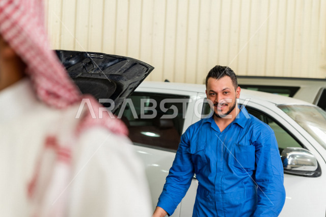 رجل  عربي سعودي يقف مع المهندس الميكانيكي داخل ورشة السيارات  ، بايماءات وجه و يدين مختلفة  ، خدمات اصلاح و صيانة السيارات ، مهندس ميكانيكي ، ورشة صيانة 
