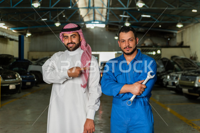 رجل  عربي سعودي يقف مع المهندس الميكانيكي داخل ورشة السيارات  ، بايماءات وجه و يدين مختلفة  ، خدمات اصلاح و صيانة السيارات ، مهندس ميكانيكي ، ورشة صيانة 