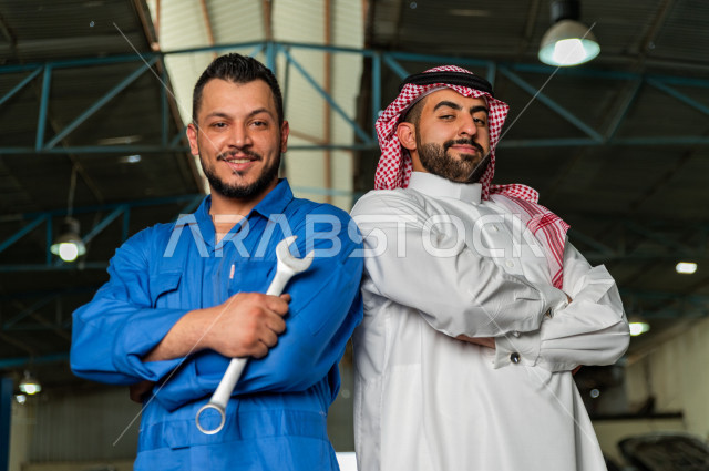 رجل  عربي سعودي يقف مع المهندس الميكانيكي داخل ورشة السيارات  ، بايماءات وجه و يدين مختلفة  ، خدمات اصلاح و صيانة السيارات ، مهندس ميكانيكي ، ورشة صيانة 